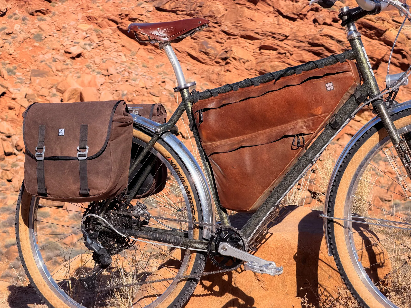 Both the waxed canvas pannier and leather framebag visible together on the bike against red desert scrub